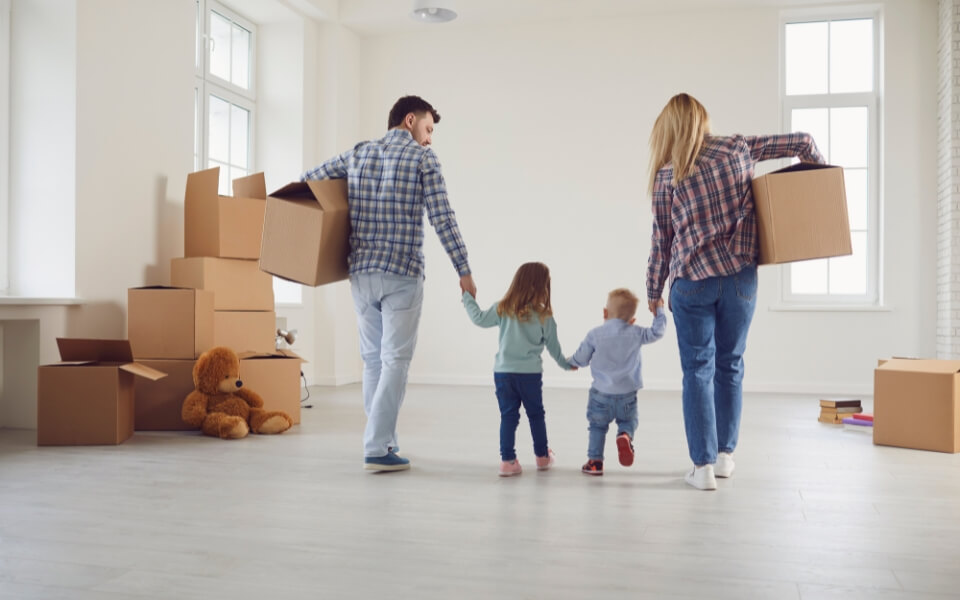 Parents with two young children, carrying moving boxes into a clean home.